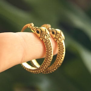 Puede incluir: Un par de pendientes de aro dorados con forma de serpiente. Las cabezas de serpiente tienen escamas detalladas y ojos negros. Los pendientes se muestran en un dedo sobre un fondo verde borroso.