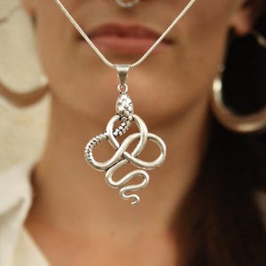 Puede incluir: Collar con colgante de serpiente plateada. El colgante presenta un diseño detallado de serpiente con cabeza y cuerpo texturizados, enrollado en un nudo intrincado. El collar se lleva en una cadena plateada.