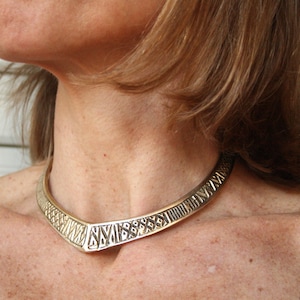 May include: A gold-toned choker necklace with a geometric pattern. The necklace is made of metal and has a V-shaped design.