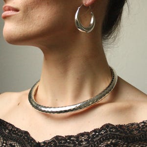 May include: A silver hammered choker necklace with a smooth, rounded shape. The necklace is worn by a person with dark hair.