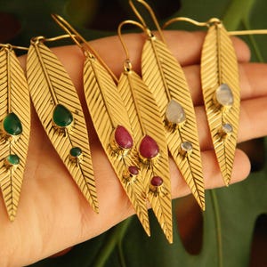 Op de afbeelding: Goudkleurige oorbellen in de vorm van een blad met edelsteenaccenten. Elke oorbel heeft een lang, getextureerd bladerontwerp met een haaksluiting. De edelstenen zijn smaragdgroen, robijnrood en maansteen.