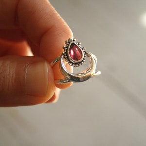 Peut inclure: Une bague en argent ornée d'une pierre rouge en forme de goutte d'eau. La bague présente un design détaillé avec une bordure perlée autour de la pierre et une monture à double bande. La bague est tenue par une main.
