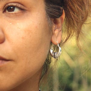 May include: Silver-toned crescent-shaped hoop earrings with a detailed band. The earrings are worn on the ear of a person. The earrings have a hook closure. The background is blurred, with natural light.