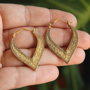 Puede incluir: Un par de pendientes de aro dorados con silueta en forma de V. Los pendientes presentan un intrincado patrón de volutas en relieve en la superficie frontal e incluyen un mecanismo de cierre de bisagra en la parte superior para sujeción.