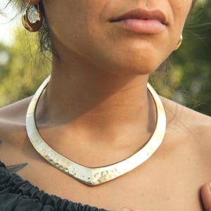 Puede incluir: Un collar de metal dorado martillado con un diseño en forma de V. El collar se lleva alrededor del cuello y se combina con pendientes de aro dorados. El fondo está borroso, lo que sugiere un entorno al aire libre.