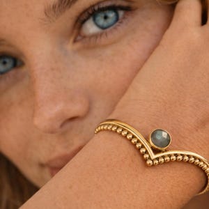 Può includere: Un braccialetto dorato con un design a forma di V e una gemma grigia rotonda al centro. Il braccialetto è adornato con piccoli dettagli a perline lungo i bordi. Il gioiello è indossato al polso.