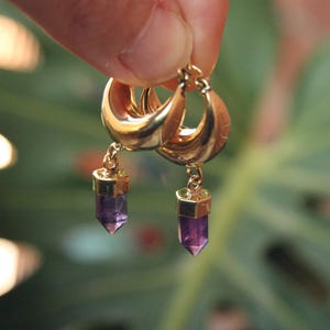 Peut inclure: Boucles d'oreilles créoles dorées avec un pendentif en pierre précieuse violette facettée. Les boucles d'oreilles sont présentées sur un fond vert flou. Elles ont une finition polie et un design simple et élégant.