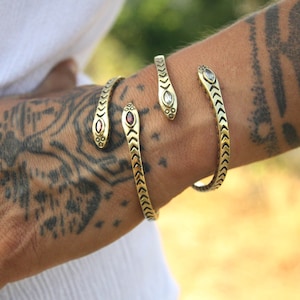 May include: Gold-coloured snake-shaped bracelets with gemstone accents. The bracelets are worn on the wrist and forearm. The snake heads feature clear and red gemstones. The bracelets have a textured, scale-like design.