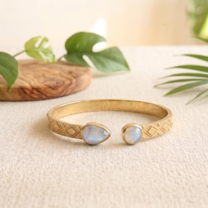 May include: A gold-colored open cuff bracelet with a geometric pattern and two light blue moonstone accents. The bracelet is displayed on a neutral surface with green foliage in the background.