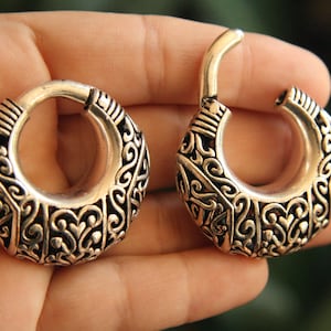 May include: A pair of silver-toned hoop earrings featuring a hinged segment and an intricate, raised scrollwork pattern across the outer surface. The earrings have a thick, crescent-shaped profile with a polished finish and dark recessed detailing.