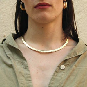 May include: A gold choker necklace with a hammered texture. The necklace is worn by a person with long brown hair and is sitting against a light beige wall.