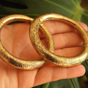 Pode incluir: Dois braceletes circulares grossos em tom dourado com um padrão intrincado de flores e vinhas gravado por toda a superfície. As pulseiras possuem um acabamento polido e reflexivo com um perfil arredondado e substancial.