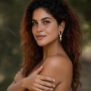 May include: A woman with auburn curly hair and freckles is wearing gold earrings and a nose ring. The earrings have a layered design. The background is a blurred green.