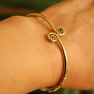 May include: A gold-coloured bracelet with a spiral design at each end. The bracelet is curved and wraps around the wrist. The metal has a shiny finish.