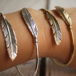 May include: Three silver and gold feather bangle bracelets on a person's wrist. The bracelets are simple and delicate, with a single feather charm on each.