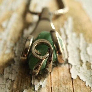 Puede incluir: Un colgante de plata con una piedra preciosa verde engastada en un diseño delicado y en espiral. El colgante está sobre una superficie de madera.