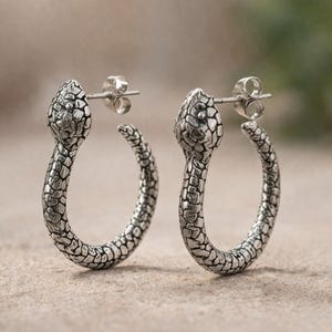 Puede incluir: Un par de pendientes de aro plateados. Cada pendiente presenta un diseño de cabeza de serpiente en la parte superior, con un cuerpo texturizado que se curva en forma de aro. Los pendientes son detallados y tienen una estética vintage.