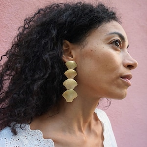 May include: A woman with long, curly black hair wearing a pair of gold, layered, fan-shaped earrings.