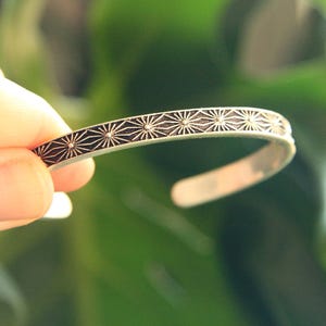 Peut inclure: Bracelet manchette en argent avec un motif géométrique répétitif en forme d'éclat d'étoile de couleur foncée. Le bracelet ouvert est présenté sur un fond vert flou, mettant en valeur le design et l'éclat métallique.