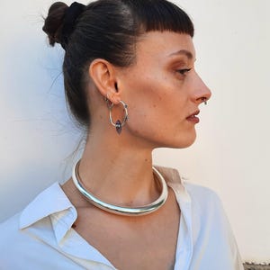 以下が含まれることがあります： 滑らかで丸みを帯びたデザインのシルバーカラーのチョーカーネックレス。画像には、宝石のアクセントが付いたフープイヤリングとノーズリングも表示されています。モデルは白い襟付きシャツを着ています。