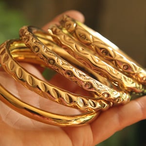 Könnte beinhalten: Ein Set aus vier goldfarbenen Metallarmreifen. Ein Armreif hat eine glatte, polierte Oberfläche, während die anderen drei verschiedene geprägte Muster aufweisen, darunter sich wiederholende ovale Vertiefungen und geometrische Gravuren.