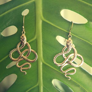 May include: A pair of gold snake earrings with a detailed, textured design. The earrings are hanging from a green leaf.