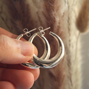 Puede incluir: Un par de pendientes de aro plateados. Los pendientes se sostienen en una mano y están enfocados. Los aros tienen un diseño simple y redondeado, con una superficie lisa y pulida. Los pendientes son un artículo de joyería clásico.