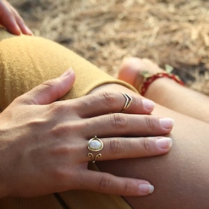 Può includere: Primo piano di una mano con due anelli d'oro. Uno presenta un motivo a chevron, l'altro una pietra rotonda e chiara in un'incastonatura filigranata. Lo sfondo è sfocato, con una gamba e un braccialetto. Estetica naturale e bohémien.