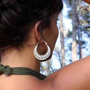 Könnte beinhalten: Eine Frau trägt einen silbernen Creolenohrring mit geometrischem Design. Der Ohrring ist aus Metall und hat ein dreieckiges Muster.