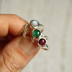 Könnte beinhalten: Drei Silberringe mit verschiedenen Edelsteinen. Der obere Ring hat einen weißen Mondstein, der mittlere Ring einen grünen Stein und der untere Ring einen roten Stein.