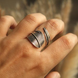 May include: A silver-toned feather ring with intricate detailing. The ring features two feather designs, one larger and one smaller, wrapping around the finger. The ring is worn on a hand, showcasing the jewelry's design and texture.