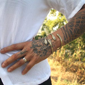May include: Gold-toned snake bracelets and a ring adorn the arm and hand. The bracelets feature snake head details. The person is wearing a white shirt. The arm has a tattoo design.