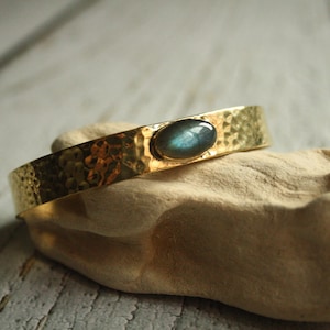 Op de afbeelding: Een gouden gegraveerde metalen armband met een blauwgroene ovale labradorit edelsteen in het midden.