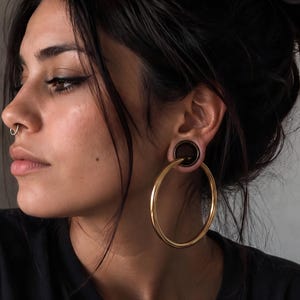 Puede incluir: Primer plano de una persona con grandes pendientes de aro dorados. Los pendientes están sujetos a tapones negros en los lóbulos de las orejas. También es visible un pequeño anillo de nariz dorado. La persona lleva una camiseta negra.