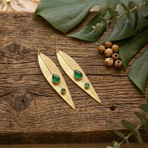 Könnte beinhalten: Goldfarbene Blatt-Ohrringe mit smaragdgrünen Edelsteinen. Die Ohrringe haben ein Federdesign und werden auf einer rustikalen Holzoberfläche mit grünem Laub und kleinen Holzperlen präsentiert. Die Ohrringe sind ein Schmuckstück.