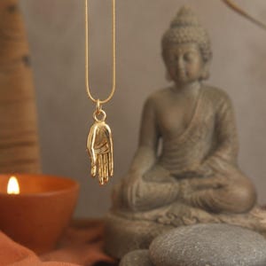 May include: A gold-colored pendant necklace featuring a detailed hand charm. The necklace hangs in front of a seated Buddha statue and a lit candle in a terracotta bowl. The background includes a neutral wall and decorative elements.