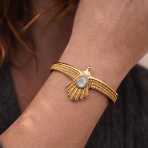Puede incluir: Brazalete dorado con un diseño de águila y una piedra central ovalada de color claro. El brazalete tiene una banda texturizada y tejida y se lleva en la muñeca. Las alas del águila están extendidas y el diseño es detallado.