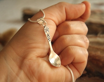 May include: A silver spoon pendant necklace. The spoon has an ornate handle design and a small, rounded bowl. The necklace chain is a delicate silver. The pendant is held in a hand.