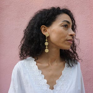 May include: A woman with long, curly dark hair wearing a white, lacy top and gold circle earrings. The earrings are a long, dangling style with multiple circles.