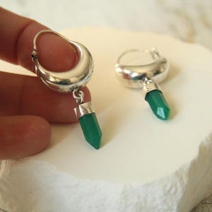 Puede incluir: Un par de pendientes de aro plateados con un acabado pulido. Cada pendiente presenta un colgante de piedra preciosa verde esmeralda facetada. Los pendientes se exhiben sobre una superficie blanca texturizada.