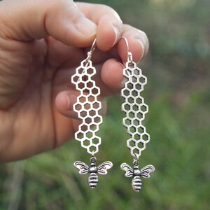 Peut inclure: Boucles d'oreilles argentées avec un motif en nid d'abeille et un pendentif abeille. Les boucles d'oreilles ont une fermeture à crochet. L'arrière-plan est flou, avec des tons verts et bruns.