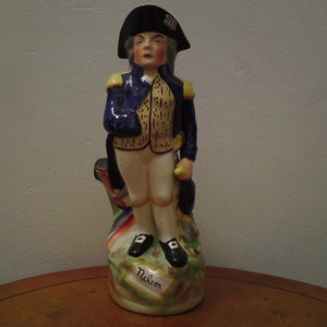 Könnte beinhalten: Eine Keramikfigur einer Person in einer blauen und goldenen Militäruniform mit einem schwarzen Hut. Die Figur steht auf einem Sockel mit der Aufschrift "Nelson".