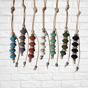 May include: A collection of handmade necklaces with lava rock beads in various colors, including blue, purple, red, white, green, and black. Each necklace features a leather cord and a silver-colored metal accent. The necklaces are displayed against a white brick wall.