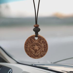 May include: A circular wooden pendant featuring a detailed carving of a wolf's face, surrounded by floral patterns. The pendant hangs from a brown cord with decorative beads. The pendant is likely a car charm or necklace.