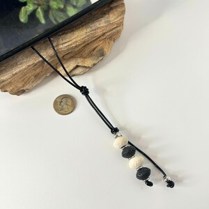 May include: Black leather phone charm with three decorative beads. The beads are a mix of cream and black, with a silver-toned metal accent. The charm is shown next to a quarter for size reference.