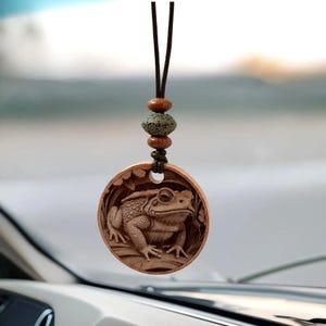 May include: A wooden frog pendant with a detailed carving of a frog inside a circular frame. The pendant hangs from a brown cord with decorative wooden and stone beads. The frog is brown and the background is blurred.