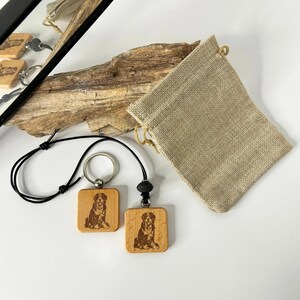 May include: Two wooden square keychains with a dog portrait, one attached to a keyring and the other to a black cord. A small beige drawstring bag and a piece of driftwood are also in the image.