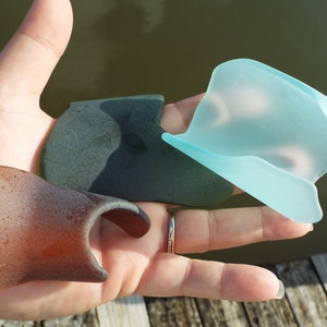 May include: Three pieces of sea glass in different colors and shapes. One piece is a dark green, one is a light blue, and one is a brown-orange.