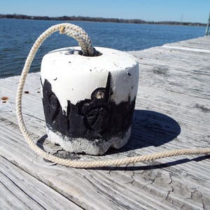 Puede incluir: Una boya cilíndrica pintada de blanco con detalles en negro y un asa de cuerda. La boya está sobre un muelle de madera desgastada cerca del agua. La cuerda es beige.