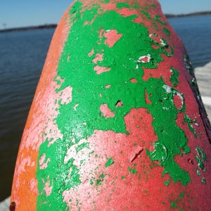 May include: A weathered, cone-shaped buoy with peeling paint. The buoy is primarily red with patches of bright green and orange. The background shows a body of water and a clear blue sky.